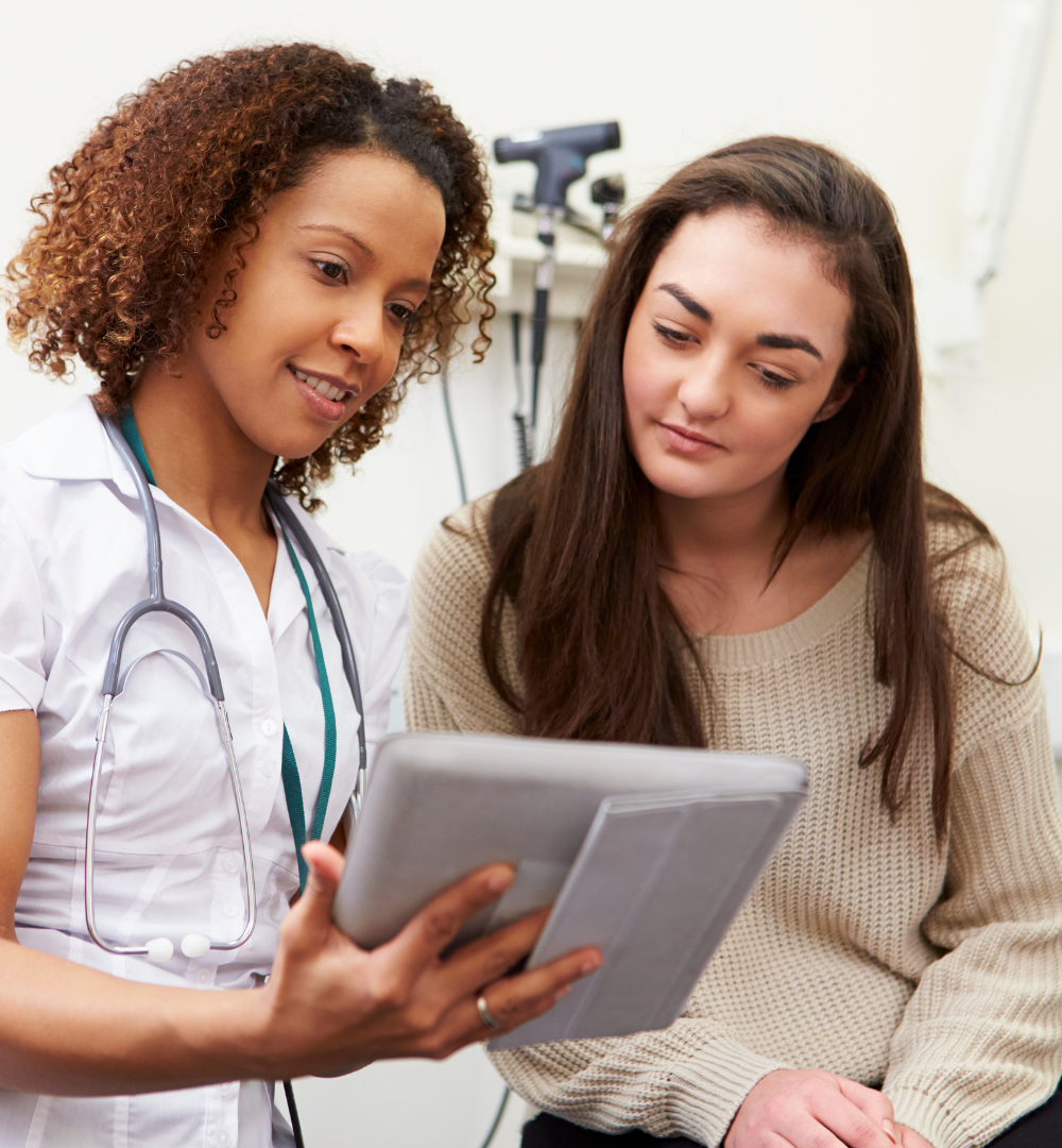 The image shows a woman in her thirties, wearing a beige shirt and looking intently at the chart her doctor is showing her. She is clearly self-advocating for her chronic illness, as she confidently reviews the healthcare plan she created with her doctor, a determined look on her face. The doctor, looking professionally attentive, is listening carefully to the woman's questions and explaining every detail. The message is clear: even with a chronic illness, self-advocacy is essential to living a full life.