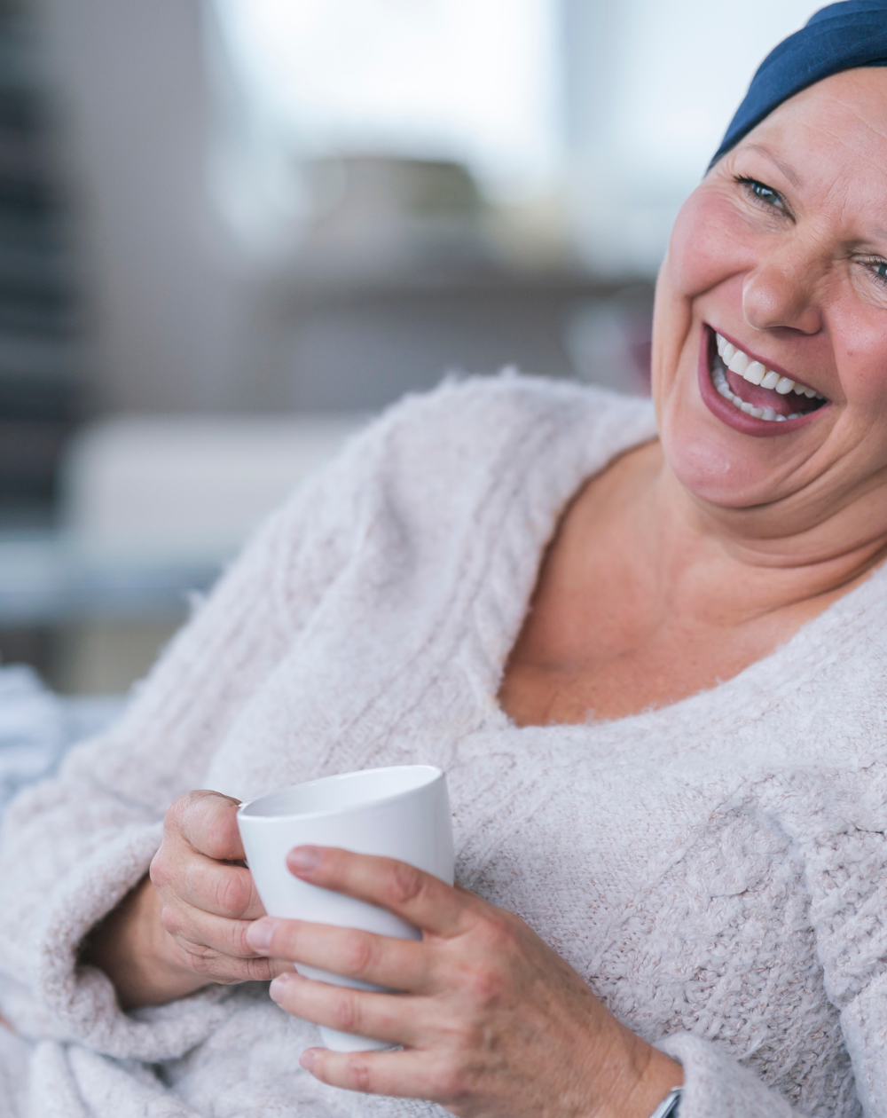 Happy woman with cancer cap on sitting on a coach holding a coffee cup and smiling.| The Suprising Benefits of Letting Your Chronic Illness Define You