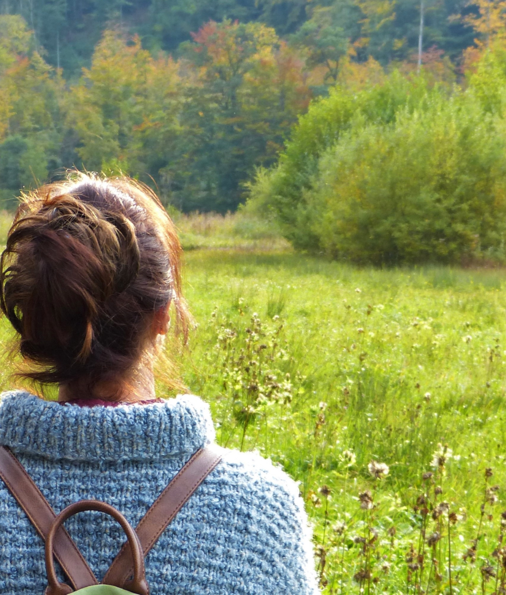 A brunette woman in a cozy grey sweater, carrying a backpack purse, gazes thoughtfully towards a serene forest path, embodying the resilience and strength discussed in the blog post about living with chronic illness. | Thriving Through the Storm: Adapting to Life With Chronic Illness